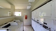 This single-floor shared laundry facility is designed with functionality and efficiency in mind. The space features a row of coin-operated dryers stacked along one wall, numbered for easy identification, alongside a separate set of top-loading washing machines. The layout is organized to allow for smooth traffic flow, with ample space to move between machines and manage laundry comfortably. A fire extinguisher is mounted on the wall, reflecting attention to safety standards within the facility. Natural light filters in through a window, brightening the otherwise utilitarian space, while overhead fluorescent lighting ensures consistent illumination throughout. The walls are finished in a neutral tone, contributing to the clean and no-frills aesthetic of the room. Informational notices are posted on the walls, providing users with relevant guidelines and usage instructions. The flooring is durable and easy to maintain, suited to the practical demands of a high-use laundry environment. Overall, this laundry facility delivers a straightforward and well-equipped space that serves the essential needs of its users efficiently.