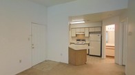 This single-floor residence offers a straightforward and functional layout designed for comfortable everyday living. The open-concept living area flows seamlessly into the kitchen, creating a connected and practical space. The living area features neutral carpeted flooring and light-toned walls, contributing to a clean and bright atmosphere. A standard entry door provides direct access to the main living space.

The kitchen is the focal point of the home, featuring cream-colored cabinetry that extends to the upper walls for ample storage. A central peninsula with a wood-toned base and a light countertop serves as a natural divider between the kitchen and living area, doubling as a breakfast bar. The kitchen is equipped with a white electric range with overhead ventilation, a full-size refrigerator, and tile flooring that contrasts with the carpeted living space. Overhead fluorescent lighting ensures the kitchen remains well-lit. The textured ceiling detail in the kitchen area adds a subtle architectural distinction between the two zones. Overall, the space presents a practical and no-frills environment well-suited for daily use.