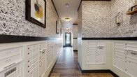 This single-floor mail room is thoughtfully designed to balance practicality with aesthetic appeal. The space is lined with an extensive array of white built-in mailbox units, systematically arranged along the walls and freestanding column partitions to accommodate a large number of residents. The mailboxes include both individual slots and larger parcel compartments, ensuring efficient mail and package management.

The design is immediately distinguished by bold, intricate black-and-white patterned wallcovering that spans the upper portions of the walls, creating a striking visual contrast against the crisp white wainscoting and mailbox cabinetry below. Dark trim accents define the transition between the two wall treatments, adding a refined, polished finish to the overall look.

Underfoot, wood-look plank flooring in warm, rich tones runs throughout the space, lending a sense of warmth and texture. Flush-mount light fixtures provide consistent, even illumination across the room. Framed artwork featuring colorful abstract compositions is displayed at regular intervals, contributing a gallery-like quality to the corridor.

The layout extends into a connecting hallway leading to an entry door, giving the space a clear and logical flow. Shelf-mounted decorative sconces add additional character. Overall, the mail room presents a cohesive, upscale aesthetic that elevates a typically utilitarian function into an engaging and well-crafted communal environment.