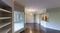 This single-floor space showcases an open-concept design that seamlessly connects the main living area to the kitchen. Rich, dark hardwood flooring runs throughout, adding warmth and a sense of continuity to the layout. The living area benefits from a large window fitted with horizontal blinds, allowing for adjustable natural light and views of the surrounding greenery outside.

Crown molding lines the ceilings, lending a polished, refined character to the interior. A pendant light fixture hangs elegantly in the transitional space between the living and kitchen areas, providing both ambient illumination and a subtle design focal point.

The kitchen is positioned to one side and features a raised bar-height counter that serves as a natural divider between the cooking and living spaces. Stainless steel appliances and a sink are visible, offering a functional and modern setup. Neutral gray walls throughout contribute to a clean, understated aesthetic that complements the dark flooring.

A hallway visible at the rear of the space leads to additional rooms, suggesting a well-organized floor plan. Ample natural light, crown molding details, and consistent finishes create a cohesive and inviting overall ambiance.