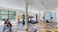 This well-appointed fitness center is designed to support a comprehensive workout experience within a bright, contemporary setting. The space is immediately distinguished by its bold, multi-colored chevron-patterned flooring in shades of yellow, purple, and gray, which adds an energetic visual character throughout.

The main exercise area is generously sized and lined with large windows that invite abundant natural light and offer views of surrounding greenery, creating an uplifting environment for cardio and strength training. Equipment includes multiple treadmills, elliptical machines, a cable weight machine, a weight rack, and a flat workout bench. Full-length mirrors along one wall visually expand the space and provide users with proper form feedback during training.

A dedicated spin and functional training studio is partitioned within the facility, housing several stationary cycling bikes, stability balls on wall-mounted racks, a balance trainer, and a ballet barre. A wall-mounted mini-split air conditioning unit ensures climate comfort in this enclosed area.

A hydration station with dual water fountains is conveniently positioned near the entrance, alongside easy access to an adjacent lounge area separated by sleek glass sliding doors. Recessed lighting throughout provides consistent, even illumination, complementing the natural light and supporting workouts at any hour.