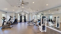 Located at 5500 Oakley Industrial Blvd in South Fulton, GA, this dedicated fitness center offers a well-appointed workout environment within a single, open-plan space. The room is defined by striking vaulted ceilings that rise to a generous peak, creating an airy and expansive atmosphere. Dual ceiling fans provide consistent airflow throughout, while recessed lighting ensures the space remains bright at all hours. Large windows along multiple walls invite abundant natural light and offer views of the surrounding greenery, enhancing the overall workout experience. The durable, rubber-composite flooring is well-suited for high-impact exercise and heavy equipment use. Cardio offerings include treadmills, elliptical trainers, and a stationary bike, while the strength training area is equipped with Paramount-brand cable machines, a multi-functional weight station, and benches for a variety of exercises. Full-length mirrors are installed along one wall, visually expanding the space and allowing users to monitor their form. An arched transom window serves as an elegant architectural focal point at one end of the room, adding character to the functional space. A secondary door provides convenient access to the exterior, completing this well-designed and fully equipped fitness facility.
