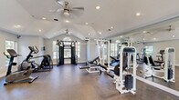 Located at 5500 Oakley Industrial Blvd in South Fulton, GA, this dedicated fitness center offers a well-appointed workout environment within a single, open-plan space. The room is defined by striking vaulted ceilings that rise to a generous peak, creating an airy and expansive atmosphere. Dual ceiling fans provide consistent airflow throughout, while recessed lighting ensures the space remains bright at all hours. Large windows along multiple walls invite abundant natural light and offer views of the surrounding greenery, enhancing the overall workout experience. The durable, rubber-composite flooring is well-suited for high-impact exercise and heavy equipment use. Cardio offerings include treadmills, elliptical trainers, and a stationary bike, while the strength training area is equipped with Paramount-brand cable machines, a multi-functional weight station, and benches for a variety of exercises. Full-length mirrors are installed along one wall, visually expanding the space and allowing users to monitor their form. An arched transom window serves as an elegant architectural focal point at one end of the room, adding character to the functional space. A secondary door provides convenient access to the exterior, completing this well-designed and fully equipped fitness facility.