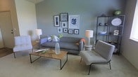 This single-floor space showcases a well-appointed living area designed with a contemporary aesthetic and a calm, inviting ambiance. The room is anchored by a soft sage-green accent wall that serves as a backdrop for a carefully curated gallery wall arrangement, featuring an eclectic mix of framed artwork in varying sizes and styles, adding visual interest and personality to the space.

The living area is furnished with a low-profile gray sofa dressed with patterned throw pillows and a pop of color from an orange accent throw. A sleek, tufted light gray accent chair complements the seating arrangement, while a wood-top coffee table with a minimalist metal frame sits at the center, styled with a glass vase of greenery and a decorative tray. Ambient lighting is provided by two matching table lamps with white shades, lending a warm glow to the room.

A slender black metal open shelving unit along one wall displays decorative objects, adding both storage and style. Neutral carpeted flooring ties the elements together, contributing to the room's relaxed yet polished atmosphere. Overall, the space feels thoughtfully designed, balancing comfort and modern style seamlessly.