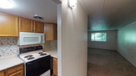 This single-story space is thoughtfully laid out across several distinct areas visible in the floor plan. The kitchen is well-equipped, featuring warm honey-toned oak cabinetry extending to the ceiling, a decorative mosaic tile backsplash, light countertops, and a full suite of white appliances including a refrigerator, electric coil range, and over-the-range microwave. Wood-look flooring runs throughout the kitchen, adding a practical and cohesive aesthetic. A partial wall separates the kitchen from the adjacent living area, which features neutral carpet flooring and a window that brings in natural light with views of surrounding greenery.

The bedrooms are carpeted and finished with white walls and paneled ceilings. Each room includes a wall-mounted HVAC unit for individual climate control, and windows fitted with horizontal blinds allow natural light while maintaining privacy. The rooms offer a clean, open canvas suitable for various furnishing arrangements.

The bathroom is finished with a white color palette throughout, featuring a tub-shower combination with a built-in shelf, a wood-framed medicine cabinet mirror, and a vanity with an integrated sink and wood cabinetry below. Wood-look flooring completes the bathroom's practical and tidy design.