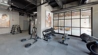 This fitness center is thoughtfully designed to support a wide range of workout routines within a single open floor layout. The space features an industrial-style aesthetic, with exposed ceiling ductwork, black-painted overhead infrastructure, and sleek linear lighting that gives the gym a modern, energetic atmosphere. Large floor-to-ceiling windows line one wall, flooding the cardio zone with natural light and offering views of the surrounding tree-lined exterior — an inviting setting for treadmill and elliptical sessions. The cardio area is well-appointed with multiple treadmills, elliptical trainers, and stationary bikes positioned to take full advantage of the natural light. The strength and free weights section features a comprehensive dumbbell rack, a multi-station cable and weight machine, adjustable benches, medicine balls, and a balance ball, with full-length mirrors along the wall to assist with form. A dedicated functional training zone includes a freestanding heavy punching bag and additional specialty equipment. Motivational fitness imagery is displayed prominently on the walls, reinforcing the gym's energetic character. Secure storage lockers and a package system are conveniently accessible within the space. The flooring throughout consists of durable interlocking carpet tiles suited for high-traffic athletic use. An adjacent lounge area, visible through glass partition walls, provides a comfortable retreat for rest and recovery.