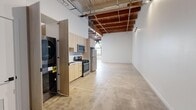 Located at 2349 S Santa Fe Ave in Los Angeles, CA, this industrial-style unit blends raw architectural character with modern finishes across a single open floor plan. The space is defined by its striking exposed wood beam ceiling, which runs the length of the unit and contributes to a loft-like ambiance with generous vertical clearance. Polished concrete floors extend throughout, reflecting light and reinforcing the clean, minimalist aesthetic.

The kitchen is well-appointed with light wood cabinetry extending to ceiling height, a white quartz countertop, a stainless steel undermount sink with a matte black faucet, and a full suite of stainless steel appliances including a gas range, microwave, dishwasher, and refrigerator. A stacked washer/dryer unit is neatly housed behind a dedicated cabinet panel adjacent to the kitchen.

The main open area offers a generously proportioned, flexible space with crisp white walls and track lighting mounted to the ceiling beams — well-suited for living, working, or creative use. Natural light filters in through a window visible at the far end of the unit. The bathroom features full white subway tile surrounding a soaking tub and shower combo, accented by matte black fixtures for a modern industrial touch.