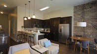 This single-floor space blends modern design with industrial charm in a thoughtfully laid-out open-concept floor plan. The kitchen is a standout feature, showcasing rich dark cabinetry that extends to the ceiling for ample storage, stainless steel appliances including a French door refrigerator with ice and water dispenser, and a built-in microwave. A light-toned countertop peninsula with pendant lighting serves as both a functional prep area and a casual dining bar with seating, making it an inviting hub for entertaining. The kitchen flows naturally into the living area, which is anchored by a comfortable seating arrangement featuring neutral-toned chairs accented with colorful throw pillows. An exposed brick accent wall on one side adds warmth and a distinctive industrial character to the space, complementing the hardwood-style flooring that runs throughout. The high ceilings contribute to an airy, open ambiance and are enhanced by a recessed ceiling light fixture that brightens the interior. A small dining nook is also present, offering an intimate setting for meals. Overall, the space combines practical functionality with a warm, contemporary aesthetic.