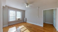 This single-floor unit offers a clean, functional layout with a cohesive and straightforward design throughout. The main living area is anchored by warm, honey-toned hardwood floors that reflect natural light pouring in through windows fitted with blinds. A ceiling fan with an integrated light fixture provides both ventilation and ambient lighting. Classic white baseboards and neutral gray-toned walls create a bright, airy feel throughout the space. A cast-iron radiator adds a charming, traditional touch to the living area, hinting at the building's heritage. The bedroom continues the same hardwood flooring and benefits from its own ceiling fan and window-mounted air conditioning unit. A spacious closet offers practical storage. The kitchen is efficiently laid out with white upper and lower cabinetry, dark speckled countertops, a stainless steel sink, and a gas range, all set atop tile flooring. The full bathroom presents a clean, all-white aesthetic with a soaking tub, pedestal sink, toilet, and a window that allows natural light to brighten the space. Tile flooring and wainscoting-style wall treatments complete the bathroom's classic look. Overall, the unit maintains a tidy, well-lit atmosphere suited for comfortable everyday living.