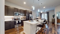 This single-floor residence is designed with a contemporary aesthetic and a thoughtful open-concept layout. The kitchen serves as a central focal point, featuring dark espresso cabinetry that extends to the ceiling, a textured tile backsplash, and a full suite of stainless steel appliances including a French door refrigerator, electric range, and over-the-range microwave. A large white quartz island with an undermount sink and seating for multiple bar stools anchors the space, with pendant lighting suspended above. Rich hardwood-style flooring flows continuously throughout the main living areas, complementing the clean white walls and recessed lighting overhead.

The living area is bright and welcoming, benefiting from natural light that pours through a set of French doors dressed with linen drapes, opening to an outdoor area. A ceiling fan adds comfort to the space.

The bedroom is carpeted and comfortably scaled, with a window allowing natural light and ample wall space. The primary bathroom is generously proportioned, offering a dual-sink vanity with dark cabinetry, hardwood-style flooring, a soaking tub, and a separate walk-in closet with built-in shelving and rod storage.