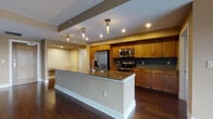 This single-floor, 1-bedroom, 1.5-bathroom residence is thoughtfully designed to maximize both function and style. Rich dark hardwood flooring flows throughout the open-concept living and kitchen areas, creating a warm and cohesive aesthetic. The kitchen is a standout feature, showcasing warm-toned wood cabinetry, a teal mosaic tile backsplash, stainless steel appliances, and a generously sized granite-topped island with pendant lighting overhead — ideal for both meal prep and casual dining. The living area is bright and airy, anchored by large sliding glass doors that open to a balcony and frame expansive outdoor views of a waterway and city skyline beyond. The spacious bedroom is carpeted for comfort, featuring a ceiling fan, large windows with the same scenic views, and access to a full en-suite bathroom with a walk-in closet. The en-suite bath is finished with tile flooring, a wood-toned vanity, and clean, modern fixtures. A convenient half bathroom, tiled with classic subway tile wainscoting and a pedestal sink, serves guests. Throughout, warm neutral wall tones and recessed lighting contribute to a welcoming, well-lit ambiance.