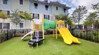 This cheerful outdoor playground is designed to provide a safe and engaging environment for children. The centerpiece of the space is a multi-component play structure finished in bright yellow and green, featuring a distinctive leaf-shaped tunnel, a yellow double slide, climbing stairs, and an interactive number activity panel. A secondary log-style slide adds additional play variety. The entire area is laid out on well-maintained artificial turf, offering a clean, cushioned surface ideal for active play. The playground is enclosed by black iron fencing, providing a secure boundary for children. Tall palm trees and carefully landscaped greenery frame the perimeter, lending a tropical character to the setting. The backdrop features a multi-story residential building with a classic coastal aesthetic, characterized by light blue and white board-and-batten siding, black shutters, and white-trimmed windows. The combination of vibrant play equipment, lush surroundings, and the architectural charm of the neighboring buildings creates an inviting and family-friendly atmosphere. The open layout of the playground ensures excellent sightlines for supervision while allowing children ample space to move freely and explore.