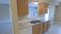 This single-story space includes a well-appointed kitchen that serves as a central feature of the home. The kitchen is outfitted with warm-toned oak wood cabinetry, offering generous upper and lower storage throughout. Countertops provide ample workspace and house a stainless steel double-basin sink, complemented by a white dishwasher positioned conveniently nearby. A notable architectural detail is the pass-through window cut into the wall, which opens the kitchen visually to an adjacent room, allowing natural sightlines and enhancing the sense of openness between spaces. The adjacent area visible through the pass-through features built-in shelving, adding functional storage and character. Light tile flooring runs through the kitchen, contributing to a clean, easy-to-maintain aesthetic. Natural light filters in from a window to the right, brightening the space and highlighting the neutral tones of the cabinetry and countertops. A flush-mount ceiling light provides consistent overhead illumination. Overall, the kitchen presents a practical and cohesive design with classic finishes suited for everyday living.