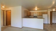 This single-floor space features an open-concept layout that connects the kitchen and living area, creating a functional and cohesive floor plan. The kitchen is equipped with light wood cabinetry offering ample storage through both upper and lower cabinets. A built-in range with an overhead ventilation hood and a full-size refrigerator provide essential appliances, while the countertops in a neutral tone complement the warm wood tones of the cabinetry. A peninsula-style counter separates the kitchen from the adjacent living area, serving as a natural dividing element while maintaining the open feel of the space. The living area is finished with wall-to-wall carpet in a light neutral tone, contributing to a soft and understated aesthetic throughout. Walls are painted in a clean, bright white, enhancing the sense of openness. Overhead flush-mount lighting provides consistent illumination across the kitchen area. The overall design is straightforward and practical, featuring a cohesive neutral palette of whites, beiges, and light wood tones that lend the space a clean, move-in-ready appearance suitable for a variety of furnishing styles.