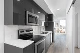 This single-floor unit presents a clean, contemporary design characterized by crisp white walls, light wood-tone flooring, and an efficient layout that maximizes functionality throughout. The galley-style kitchen is a standout feature, outfitted with dark cabinetry contrasted against light stone countertops, a subway tile backsplash, and a full suite of stainless steel appliances, including a refrigerator, electric range, and over-the-range microwave. Recessed lighting illuminates the kitchen corridor, which connects seamlessly to the main living area. The open living space benefits from expansive floor-to-ceiling windows that flood the interior with abundant natural light while framing impressive views of surrounding buildings, greenery, and a body of water beyond. A secondary room, accessible via a sliding door, offers additional flexibility and similarly enjoys natural light through its own window with urban views. The bathroom is simply appointed with light-toned finishes and wood-look flooring consistent with the rest of the unit. Throughout, the design maintains a minimalist aesthetic, with neutral tones and clean lines creating a bright, cohesive atmosphere well-suited to modern living.