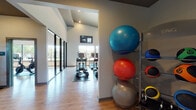 Located at 4600 N Josey Ln in Carrollton, Texas, this well-appointed fitness center is thoughtfully organized across a single floor into distinct workout zones, each designed to support a comprehensive exercise experience. The cardio area is generously sized, housing a row of TRUE-brand treadmills and elliptical trainers positioned beneath soaring vaulted ceilings. Multiple wall-mounted flat-screen displays stream workout content, while large windows allow ample natural light to fill the space and offer views of the surrounding landscape. A dedicated cycling studio features a fleet of Peloton and other stationary bikes arranged on wood-style flooring, with an accent wall in bold teal, a wall-mounted screen, and wide windows that bring in outdoor views. The strength training zone is equipped with a dumbbell rack offering a full range of free weights, a weight bench, and cable/resistance machines, complemented by high clerestory windows that maintain brightness without compromising wall space. Additional equipment includes a rowing machine, plyometric jump box, and a rack stocked with stability balls and kettlebells. Throughout the facility, rubber mat flooring protects workout areas, and the consistent use of neutral tones with clean architectural lines creates a focused, energizing environment suited for a full range of fitness activities.