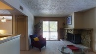 This single-floor space offers a comfortable and functional layout centered around a welcoming living area. The room is anchored by a striking stone fireplace, which serves as a natural focal point and adds a rustic, cozy character to the space. Adjacent to the fireplace, a sliding glass door opens to a private balcony, flooding the interior with natural light and providing pleasant views of the surrounding greenery and trees. The textured ceiling adds visual depth and a classic residential feel throughout. Soft carpeted flooring contributes to the warm ambiance, complementing the neutral, earthy wall tones. A louvered closet door is visible, offering practical storage solutions. Décor elements such as framed artwork and indoor plants enhance the lived-in, inviting quality of the space. A glass-top coffee table and seating area complete the living space, making it well-suited for relaxation and everyday comfort. Overall, the space balances functional design with a warm, homey aesthetic, ideal for comfortable single-level living.