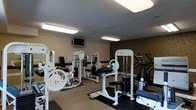 This fitness center offers a practical and well-organized workout environment designed to accommodate a variety of exercise routines. The space is outfitted with multiple white-framed strength-training machines, including cable systems, a leg extension/curl station, and a chest/shoulder press, all positioned on protective rubber flooring mats to ensure safety and stability during use. Cardio equipment, including treadmills, is arranged along the far wall, providing users with a full range of exercise options. A wall-mounted flat-screen television is installed for entertainment during workouts, adding to the overall comfort of the space. The room is finished with neutral-toned walls featuring a subtle textured lower border, contributing to a clean and focused atmosphere. Overhead fluorescent lighting ensures bright, even illumination throughout the entire area, making it easy to navigate the equipment at any time of day. The layout is thoughtfully arranged to maximize available floor space while allowing for safe movement between stations. Overall, this fitness center provides a functional and well-maintained environment suited for both strength training and cardiovascular workouts.