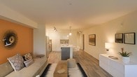 This single-floor space showcases a thoughtfully designed open-concept layout that connects the living, dining, and kitchen areas into one cohesive and functional environment. Light wood-tone flooring extends throughout, lending warmth and visual continuity to the space.

The kitchen is outfitted with sleek white cabinetry reaching toward the ceiling, stainless steel appliances including a refrigerator and built-in oven, and a central island with a contrasting dark base. The island doubles as a casual dining area, accommodating bar-style seating. Pendant lighting above the island and track lighting in the kitchen provide focused task illumination, complementing the overall warm ambient lighting.

The living area is generously proportioned, featuring a comfortable seating arrangement anchored by a patterned area rug and a low-profile coffee table. A framed landscape artwork adds a focal point to the main wall, enhancing the refined yet understated aesthetic.

A hallway visible in the background suggests access to additional private areas of the unit. The overall ambiance is clean, contemporary, and inviting, with a neutral palette that allows natural and artificial light to enhance the sense of openness.