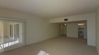 This single-floor space offers a clean, understated layout designed for comfortable everyday living. The main living area is generously proportioned, featuring wall-to-wall carpet in a warm neutral tone that runs throughout, contributing to a cohesive and calm aesthetic. Smooth, light-colored walls and a flat white ceiling create a bright, airy atmosphere, while a HVAC vent integrated into the ceiling ensures year-round climate comfort. Vertical blinds on one side of the room allow for adjustable natural light, casting soft rays across the floor and adding to the space's welcoming feel. A subtle step-down or architectural ledge separates the main living area from an adjoining section, adding a touch of structural interest and visual definition to the open floor plan. A hallway visible from the main room leads to additional spaces, suggesting a practical and functional layout. Overhead lighting in the hallway provides a well-lit transition between areas. The overall ambiance is neutral and move-in ready, offering a versatile canvas well-suited to a variety of interior design preferences. The single-floor arrangement ensures ease of navigation throughout the space.