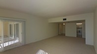 This single-floor space offers a clean, understated layout designed for comfortable everyday living. The main living area is generously proportioned, featuring wall-to-wall carpet in a warm neutral tone that runs throughout, contributing to a cohesive and calm aesthetic. Smooth, light-colored walls and a flat white ceiling create a bright, airy atmosphere, while a HVAC vent integrated into the ceiling ensures year-round climate comfort. Vertical blinds on one side of the room allow for adjustable natural light, casting soft rays across the floor and adding to the space's welcoming feel. A subtle step-down or architectural ledge separates the main living area from an adjoining section, adding a touch of structural interest and visual definition to the open floor plan. A hallway visible from the main room leads to additional spaces, suggesting a practical and functional layout. Overhead lighting in the hallway provides a well-lit transition between areas. The overall ambiance is neutral and move-in ready, offering a versatile canvas well-suited to a variety of interior design preferences. The single-floor arrangement ensures ease of navigation throughout the space.