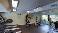 Located at 1933 North Hills Dr in Raleigh, NC, this fitness center offers a dedicated, single-floor workout facility designed to support a well-rounded exercise routine. The space is outfitted with a variety of equipment to accommodate different training styles, including a multi-function cable and weight machine for strength training, a row of treadmills for cardio, a medicine ball rack stocked with multiple weighted balls, and a dedicated bench press station. The dark rubberized flooring provides a durable, slip-resistant surface well-suited for high-activity use. Overhead, rows of fluorescent lighting ensure the space is brightly and evenly illuminated throughout. The light-colored walls keep the interior feeling open and clean, while a window along one wall introduces natural light and a view of the surrounding greenery. Posted fitness area rules and reference charts are visible on the walls, reinforcing a structured and informative environment. The layout is thoughtfully organized to allow users to move freely between equipment zones without congestion, making this fitness center a practical and functional amenity for residents at this Raleigh location.