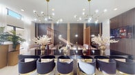 This elegantly designed single-floor space blends modern glamour with refined comfort across a thoughtfully appointed open-plan layout. Large format white porcelain tile floors and crisp white walls create a luminous backdrop, enhanced by abundant natural light streaming through floor-to-ceiling windows and clerestory transom windows positioned high along the perimeter. Sheer white drapery adds a soft, airy quality throughout.

The main gathering area features a striking central bar or lounge table lined with plush navy velvet barrel chairs accented with gold bases, illuminated overhead by sculptural brass-and-globe pendant chandeliers. A dedicated private dining or meeting nook is defined by rich dark wood wall paneling and decorative lattice screens with intricate geometric patterns, offering intimate booth-style seating with upholstered banquettes and wood-topped tables.

A casual lounge zone provides comfortable sectional seating alongside reclaimed wood accent tables, with direct sightlines to the outdoor terrace through expansive glass doors. The refreshment bar area includes a clean white countertop, built-in dark cabinetry, a sink, and globe pendant lighting, complemented by a full-length mirror wall that amplifies the sense of space. Glass double doors open onto an outdoor terrace, seamlessly connecting the interior to an al fresco setting.
