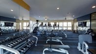 This fitness center is designed to support a comprehensive workout experience within a single, open-plan floor. The space is bright and airy, benefiting from a generous row of windows along the perimeter walls that allow abundant natural light to fill the room. Ceiling fans are evenly distributed throughout, ensuring proper air circulation during workouts.

The cardio section is prominently featured, housing a variety of machines including treadmills, ellipticals, stair climbers, and stationary bikes, all arranged to maximize space and usability. A full-length mirror lines one wall, enhancing the sense of openness and allowing users to monitor their form during exercise.

The free weights area is well-stocked with a wide range of dumbbells neatly organized on tiered racks, accompanied by weight benches suited for various strength training exercises. Exercise balls are also available throughout the space, adding versatility to any workout routine. A dedicated wall-mounted rack holds medicine balls, catering to functional fitness needs.

The flooring features durable, dark carpet tiles designed to withstand heavy equipment and high foot traffic. Recessed lighting supplements the natural light, ensuring the space remains well-lit at all hours. The overall ambiance is clean, organized, and purposefully designed to accommodate a wide range of fitness activities.