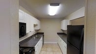 This single-floor space centers around a well-organized galley-style kitchen designed for efficient use of space. The kitchen features white shaker-style cabinetry lining both walls, providing ample upper and lower storage throughout. Dark speckled countertops run along each side, offering generous workspace for meal preparation. Black appliances, including a refrigerator, electric range, microwave, and dishwasher, are seamlessly integrated into the layout, creating a cohesive color contrast against the white cabinetry. A double-basin sink with a single-handle faucet is positioned along one wall beneath the upper cabinets. The light tile flooring brightens the space and complements the overall color palette. Overhead, a flush-mount ceiling light fixture provides central illumination, while a textured ceiling adds subtle visual interest. The neutral-toned walls and clean lines give the kitchen a tidy, functional appearance. The galley configuration makes efficient use of the available space, ensuring that storage, prep areas, and appliances are all within easy reach, making the kitchen practical for everyday use.