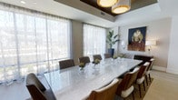 This single-floor space showcases a refined dining area that serves as its centerpiece. Dominating the room is a generously sized marble-top dining table with a striking white veined surface, surrounded by a mix of upholstered chairs in neutral tones and rich wood-framed seating, offering both comfort and style. The table is tastefully dressed with glass vase centerpieces holding protea blooms, adding a touch of organic elegance.

Overhead, a coffered ceiling detail in a deep wood tone frames a pair of contemporary drum pendant lights, creating a warm and focused ambiance above the table. Large windows dressed in sheer, patterned curtains line one wall, allowing generous natural light to filter softly into the space while maintaining privacy.

A tall potted tropical plant adds a natural, sculptural element to the corner, while a bold figurative painting on the adjacent wall introduces a vibrant artistic accent. A sideboard visible in the background provides functional storage and display space for glassware and accessories. The overall aesthetic is polished and contemporary, blending warm materials with clean architectural lines.