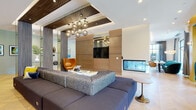 This thoughtfully designed amenity floor delivers a diverse collection of social, recreational, and productive spaces, all united by a bold contemporary aesthetic. The central lounge impresses with a textured ceiling featuring metallic mesh panels, a cluster of sculptural globe pendant lights, warm wood-paneled walls, and a sleek see-through fireplace that anchors the room. Adjacent to the lounge, a co-working area offers glass-enclosed private work pods framed by mirrored, textured columns, providing focused workspaces alongside casual lounge seating. The café-style dining area is bathed in natural light through floor-to-ceiling windows and glass doors that open to an outdoor patio strung with café lights. Light herringbone hardwood flooring flows throughout, complementing the warm amber and teal color palette. A dedicated games room features a billiards table and ping pong, enhanced by dramatic cove lighting and textured accent walls. The golf simulator room provides an immersive experience with a full-wall projection screen. Cozy window nooks with bold teal lounge chairs and a banquette seating area backed by vertical wood slat panels round out a space designed equally for relaxation, collaboration, and entertainment.