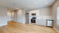 This single-floor unit at 1600–1638 State Line Rd in Kansas City, Kansas, offers a clean, modern design with a thoughtfully organized layout. Light-toned hardwood flooring extends throughout the entire space, lending warmth and visual continuity from room to room. The open-plan living and kitchen area forms the heart of the unit, with natural light filtering in through double windows fitted with white horizontal blinds. The kitchen is well-appointed with stainless steel appliances, including a refrigerator, electric range, built-in microwave, and dishwasher. White flat-panel cabinetry provides ample storage, while a white subway tile backsplash adds a crisp, contemporary accent above the countertops. Recessed lighting overhead keeps the kitchen bright and functional. Two hallways connect the main living space to the remaining rooms, offering a sense of flow and privacy. The bathroom features a modern vanity with an integrated sink, large wall mirror, a glass-enclosed shower, and consistent light wood flooring that ties it to the rest of the unit. Neutral gray walls throughout contribute to a calm, cohesive aesthetic. The overall design is understated yet functional, making efficient use of the available footprint.