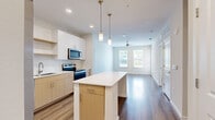This contemporary single-floor residence showcases a thoughtfully designed open-concept layout that seamlessly connects the kitchen and living areas. The kitchen is a standout feature, equipped with stainless steel appliances including a French door refrigerator with ice maker, electric range, built-in microwave, and dishwasher. A central island with a clean white countertop provides additional prep and dining space, while dual-tone cabinetry — combining white upper cabinets with warm wood-toned lower cabinets — adds modern visual depth. White tile backsplash and recessed lighting complete the polished look. Elegant pendant lights with brass accents hang above the island, enhancing the contemporary aesthetic. The living area extends naturally from the kitchen, bathed in natural light from large windows and outfitted with a ceiling fan. Wood-style flooring runs consistently throughout the residence, lending warmth and cohesion to every room. The bedrooms are bright and airy, each featuring multiple windows with blinds that invite natural light and outdoor views. The bathroom includes a soaking tub and clean white fixtures. With neutral wall tones, clean lines, and an abundance of natural light, this residence offers a fresh, move-in-ready environment with a modern sensibility.