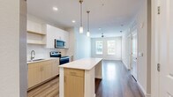 This contemporary single-floor residence showcases a thoughtfully designed open-concept layout that seamlessly connects the kitchen and living areas. The kitchen is a standout feature, equipped with stainless steel appliances including a French door refrigerator with ice maker, electric range, built-in microwave, and dishwasher. A central island with a clean white countertop provides additional prep and dining space, while dual-tone cabinetry — combining white upper cabinets with warm wood-toned lower cabinets — adds modern visual depth. White tile backsplash and recessed lighting complete the polished look. Elegant pendant lights with brass accents hang above the island, enhancing the contemporary aesthetic. The living area extends naturally from the kitchen, bathed in natural light from large windows and outfitted with a ceiling fan. Wood-style flooring runs consistently throughout the residence, lending warmth and cohesion to every room. The bedrooms are bright and airy, each featuring multiple windows with blinds that invite natural light and outdoor views. The bathroom includes a soaking tub and clean white fixtures. With neutral wall tones, clean lines, and an abundance of natural light, this residence offers a fresh, move-in-ready environment with a modern sensibility.