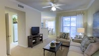 This single-story residence offers a well-appointed living area designed for both comfort and functionality. The living room is finished with warm neutral-toned walls, crown molding, and plush carpeting, creating a welcoming and cohesive aesthetic. A ceiling fan with a built-in light fixture anchors the room, providing both airflow and ambient lighting. Large windows dressed with blinds and sheer curtains allow generous natural light to fill the space while offering a glimpse of lush greenery outdoors. The room comfortably accommodates a seating arrangement centered around a dark-finished coffee table, complemented by neutral upholstered furniture and decorative accent pillows. A rustic wood media console provides practical storage and display space. An open pass-through connects the living area to an adjacent dining or kitchen space, enhancing the sense of openness throughout. A pendant light fixture is visible in the adjoining area, adding a decorative touch. The overall layout promotes a natural flow between spaces, making this residence feel airy, bright, and thoughtfully designed for everyday living.