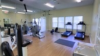 This single-floor fitness center is designed to support a well-rounded workout experience. The space is outfitted with a variety of cardio equipment, including treadmills, an elliptical trainer, and a rowing machine, providing users with multiple options for aerobic exercise. Large windows with blinds line the walls, allowing ample natural light to fill the room while maintaining privacy. The warm-toned hardwood flooring adds a clean, polished aesthetic that complements the light yellow walls. A full-length mirror spans one wall, enhancing the sense of space and allowing users to monitor their form during workouts. Overhead fluorescent lighting ensures the room is well-lit at all times of day. Exercise mats are available on the floor for stretching or floor-based routines. The layout is thoughtfully organized to allow comfortable movement between equipment. Ventilation is supported by ceiling vents, contributing to a comfortable environment during use. Overall, the fitness center presents a bright, welcoming, and functional atmosphere suited for daily exercise routines.