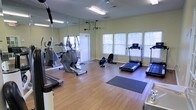 This single-floor fitness center is designed to support a well-rounded workout experience. The space is outfitted with a variety of cardio equipment, including treadmills, an elliptical trainer, and a rowing machine, providing users with multiple options for aerobic exercise. Large windows with blinds line the walls, allowing ample natural light to fill the room while maintaining privacy. The warm-toned hardwood flooring adds a clean, polished aesthetic that complements the light yellow walls. A full-length mirror spans one wall, enhancing the sense of space and allowing users to monitor their form during workouts. Overhead fluorescent lighting ensures the room is well-lit at all times of day. Exercise mats are available on the floor for stretching or floor-based routines. The layout is thoughtfully organized to allow comfortable movement between equipment. Ventilation is supported by ceiling vents, contributing to a comfortable environment during use. Overall, the fitness center presents a bright, welcoming, and functional atmosphere suited for daily exercise routines.