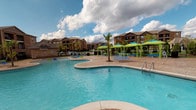 This residential community offers a thoughtfully designed set of amenities centered around comfort and active living. The outdoor area is anchored by a generously sized, freeform swimming pool with crystal-clear blue water, surrounded by a stamped concrete sun deck. The deck is furnished with lounge chairs and shaded by vibrant lime-green umbrellas and canopies, creating a resort-like atmosphere. Tall palm trees add a tropical aesthetic, while multi-story brick buildings frame the perimeter, providing an enclosed and private feel. A covered pavilion area adjoins the pool, offering a shaded gathering space.

The fitness center is divided into functional zones, decorated with bold red accent walls and rubber flooring throughout. The cardio section features a row of treadmills equipped with individual screens, positioned beneath wall-mounted televisions and contemporary artwork. The strength training area includes a multi-station cable machine, a full dumbbell rack, a weight bench, and additional resistance equipment. Large windows allow natural light to fill the space, and floor-to-ceiling mirrors enhance the sense of openness. The facility also includes an open mezzanine-style layout with railing, offering views between workout areas and creating a dynamic, energetic environment.