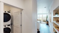 This thoughtfully designed single-floor residence offers a clean, contemporary aesthetic throughout. The open-concept layout connects the kitchen, dining, and living areas seamlessly, creating a bright and cohesive living environment. Large windows flood the interior with natural light and frame panoramic views of the surrounding treetops and urban landscape beyond.

The kitchen is well-appointed with stainless steel appliances, including a French door refrigerator, electric range, and over-the-range microwave. Sleek wood-toned upper cabinetry pairs with white lower cabinets, complemented by a white subway tile backsplash and light countertops. A set of built-in open shelves adds functional display space nearby.

The living area is generous and airy, anchored by light hardwood-style flooring that runs throughout the unit. A louvered closet door near the entry adds a classic touch, while the overall palette of white walls and neutral finishes lends the space a calm, modern character.

The bedroom is a serene retreat, featuring a large picture window with the same expansive outdoor views. The second bedroom is bright and simply finished, with its own window looking out to the greenery. Both rooms benefit from the unit's elevated position, enhancing the sense of openness and connection to the outdoors.