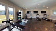 This fitness center is a thoughtfully designed, single-floor facility built to support a comprehensive workout routine. The space is finished with durable diamond-plate rubber flooring throughout, providing both safety and a clean, professional aesthetic. Recessed lighting illuminates the room evenly, complemented by generous natural light streaming through multiple large windows and glass-panel doors along the perimeter walls. These doors offer views of the surrounding outdoor areas, including a pool, adding an open, airy feel to the space.

The facility is divided into distinct workout zones. A dedicated cardio area houses multiple treadmills and an elliptical trainer, each accompanied by individual tablet or monitor displays. Multiple large flat-screen TVs are mounted along the walls, ensuring visibility from nearly every position in the room. A strength training zone features a multi-station cable machine, adjustable benches, and a rack of dumbbells and weighted medicine balls, accommodating a range of resistance exercises.

A built-in counter with a pass-through window serves as an informational station, displaying fitness center rules and guidelines. A water fountain is also conveniently accessible within the space. The neutral wall tones and clean finishes contribute to a bright, welcoming atmosphere suited for residents seeking a functional and well-maintained fitness environment.