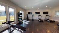 This fitness center is a thoughtfully designed, single-floor facility built to support a comprehensive workout routine. The space is finished with durable diamond-plate rubber flooring throughout, providing both safety and a clean, professional aesthetic. Recessed lighting illuminates the room evenly, complemented by generous natural light streaming through multiple large windows and glass-panel doors along the perimeter walls. These doors offer views of the surrounding outdoor areas, including a pool, adding an open, airy feel to the space.

The facility is divided into distinct workout zones. A dedicated cardio area houses multiple treadmills and an elliptical trainer, each accompanied by individual tablet or monitor displays. Multiple large flat-screen TVs are mounted along the walls, ensuring visibility from nearly every position in the room. A strength training zone features a multi-station cable machine, adjustable benches, and a rack of dumbbells and weighted medicine balls, accommodating a range of resistance exercises.

A built-in counter with a pass-through window serves as an informational station, displaying fitness center rules and guidelines. A water fountain is also conveniently accessible within the space. The neutral wall tones and clean finishes contribute to a bright, welcoming atmosphere suited for residents seeking a functional and well-maintained fitness environment.