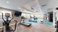 This fitness center offers a thoughtfully designed workout environment across a single level, combining functionality with a clean, contemporary aesthetic. The space is anchored by a distinctive tray ceiling with recessed lighting and dual ceiling fans, ensuring consistent air circulation throughout the room.

The flooring is a standout design element, featuring a bold geometric pattern of interlocking hexagonal tiles in shades of teal, aqua, and neutral beige, adding visual energy to the space. Multiple windows line the walls, inviting ample natural light and offering outdoor views that contribute to an open, airy atmosphere.

Cardio equipment is prominently featured, including several Matrix treadmills, an elliptical trainer, and spinning bikes. A wall-mounted flat-screen television accompanies the cardio area for entertainment during workouts. A dedicated strength training zone includes a multi-function cable machine flanked by floor-to-ceiling mirrors, a weight bench, and a dumbbell rack with a full set of free weights.

Built-in cabinetry and shelving along one wall provide practical storage, while the mirrored wall expands the sense of space. The overall ambiance is bright, clean, and inviting, creating a motivating environment well-suited for a full range of fitness activities.