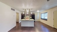 Located at 4805 Marquetter NE in Albuquerque, New Mexico, this single-floor residence offers a practical and modern layout designed for comfortable everyday living. The open-concept design seamlessly connects the kitchen and living areas, creating a spacious and functional flow throughout the main living space. The kitchen is a standout feature, equipped with dark espresso cabinetry, a black refrigerator, a built-in microwave, and ample counter space. A peninsula-style breakfast bar with a light countertop serves as both a workspace and a casual dining area, anchored by a trio of pendant lights that add a contemporary touch. The kitchen also includes a dedicated sink area and ceiling-mounted light fixtures that ensure the space is well-lit. Rich, dark hardwood-style flooring extends throughout the open living area, providing warmth and visual continuity. The living area benefits from a window that draws in natural light, enhancing the overall brightness of the space. A textured ceiling adds architectural character throughout. A hallway visible from the main living area leads to additional rooms, and a thermostat is conveniently positioned near the entry. Central HVAC is indicated by visible venting, ensuring year-round climate comfort.