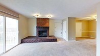 This single-floor residence offers a comfortable and well-appointed layout designed for easy living. The centerpiece of the main living area is a striking full-height brick fireplace, featuring a raised brick hearth that adds both character and warmth to the space. The fireplace's rich, dark brick tones create a bold focal point against the neutral, light-toned walls that run throughout the home. Plush wall-to-wall carpeting in a warm beige tone covers the living area floor, contributing to a cozy and inviting atmosphere. Flush-mount ceiling light fixtures provide consistent overhead illumination across the space. The living area is generously sized, offering ample room for various furniture arrangements. A white interior door adjacent to the fireplace provides access to an additional room or storage area, while an open doorway on the far side of the space connects to what appears to be a continuation of the home's interior. The overall design reflects a classic, functional aesthetic with clean lines, understated finishes, and a cohesive neutral palette that provides a versatile backdrop for personalization.