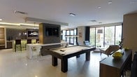 This single-floor space is designed as a contemporary entertainment and social hub, combining multiple functional zones within an open-plan layout. At the center of the room stands a dark-framed pool table with a neutral felt surface, positioned beneath a sleek rectangular pendant light that serves as a striking focal point. The surrounding floor is finished in large-format light-toned tiles, lending a clean, polished aesthetic throughout.

To one side, a bar and lounge area is anchored by a wall-mounted flat-screen television set within a dark, wood-paneled feature wall. The bar counter features modern pendant lighting and white seating, creating a refined gathering spot. Adjacent to the bar, the kitchen or service area is visible, outfitted with contemporary cabinetry and fixtures.

The opposite side of the space opens into a relaxed seating lounge with cushioned chairs positioned to take advantage of the expansive floor-to-ceiling windows and glass sliding doors. These openings flood the interior with natural light and offer direct views of a lush outdoor area, seamlessly connecting the interior to the exterior. The overall ambiance is modern, sophisticated, and well-suited for social gatherings.