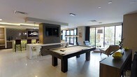This single-floor space is designed as a contemporary entertainment and social hub, combining multiple functional zones within an open-plan layout. At the center of the room stands a dark-framed pool table with a neutral felt surface, positioned beneath a sleek rectangular pendant light that serves as a striking focal point. The surrounding floor is finished in large-format light-toned tiles, lending a clean, polished aesthetic throughout.

To one side, a bar and lounge area is anchored by a wall-mounted flat-screen television set within a dark, wood-paneled feature wall. The bar counter features modern pendant lighting and white seating, creating a refined gathering spot. Adjacent to the bar, the kitchen or service area is visible, outfitted with contemporary cabinetry and fixtures.

The opposite side of the space opens into a relaxed seating lounge with cushioned chairs positioned to take advantage of the expansive floor-to-ceiling windows and glass sliding doors. These openings flood the interior with natural light and offer direct views of a lush outdoor area, seamlessly connecting the interior to the exterior. The overall ambiance is modern, sophisticated, and well-suited for social gatherings.