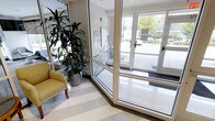 This single-story space presents a well-appointed building lobby that combines functionality with an inviting atmosphere. The entrance is defined by expansive floor-to-ceiling glass doors and sidelights, flooding the interior with natural light and providing clear views of the surrounding outdoor streetscape and landscaping.

The flooring consists of large-format polished light-toned tiles, arranged in a subtle pattern that adds visual interest while maintaining a clean, professional aesthetic. A wall-mounted intercom or directory panel is positioned near the entrance, serving as a practical wayfinding feature for visitors.

The lobby is accented with thoughtful decorative elements, including a generously sized potted tropical plant in a polished metallic planter, adding a touch of greenery and warmth to the space. A yellow upholstered accent chair provides a comfortable seating option, contributing a pop of color against the neutral tones of the walls and floors.

The exposed brick detail on one wall introduces a subtle textural contrast, complementing the otherwise contemporary finish palette. Overall, the space conveys a clean, modern, and welcoming character suited for a professional or residential building entrance.