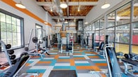 This dynamic fitness facility makes a bold first impression with its energetic design and spacious, open layout. The single-floor space features soaring ceilings with exposed wooden beams and industrial-style ductwork, lending the gym an open, airy atmosphere. Pendant lighting hangs throughout, providing warm, even illumination across the workout floor.

The flooring is one of the most striking design elements — a vibrant checkerboard pattern of orange, teal, gray, and white tiles that brings a lively energy to the space. The walls are accented with bold orange and white diagonal stripes, reinforcing the facility's spirited aesthetic.

The gym is comprehensively outfitted with a wide array of strength training equipment, including cable machines, multi-functional weight stations, benches, and free weights, all arranged to allow comfortable movement between stations. Large windows along the perimeter allow natural light to filter in, contributing to an open and inviting workout environment.

A mezzanine-level structure is visible toward the rear of the space, adding vertical dimension and additional functional areas. The overall ambiance is motivating and modern, combining industrial architectural character with a colorful, high-energy design that encourages an active and focused fitness experience.
