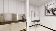 This 140 square foot pet spa room is a specialized space designed for animal grooming and care. The room features clean, white vertical paneling on the walls, creating a bright and hygienic atmosphere. A stainless steel grooming tub is prominently placed, ideal for bathing pets of various sizes. Adjacent to the tub is a professional grooming table equipped with an adjustable arm and restraints for secure pet handling. The room's practical design includes a wooden door with a glass panel, allowing visibility while maintaining a controlled environment. A framed dog portrait and a potted plant add aesthetic touches to the otherwise utilitarian space. The floor appears to be a durable, easy-to-clean material suitable for a high-traffic pet care area. Lighting fixtures, though not visible, likely provide ample illumination for detailed grooming work. This compact yet efficient layout maximizes the use of space, accommodating all necessary equipment for a full-service pet spa within its limited square footage.