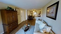 This single-floor residence offers a cohesive and functional layout that seamlessly connects the main living areas. The living room serves as the heart of the home, featuring rich hardwood flooring, warm yellow-toned walls, and a comfortable seating arrangement centered around a glass-top oval coffee table. A neutral area rug anchors the space, complementing the light-toned sofa adorned with accent pillows. Adjacent to the living room, the dining area accommodates a full dining table and chairs beneath a ceiling fan with integrated lighting, providing both comfort and practicality. The open sightlines between the living and dining spaces create an airy, connected atmosphere ideal for everyday living and entertaining. Beyond the dining area, a pass-through counter connects to the kitchen, which features cabinetry and a refrigerator visible in the background, offering a convenient and efficient cooking space. The entryway, visible along the left wall, provides a welcoming transition into the home alongside a large wooden armoire that adds both storage and character. Framed artwork adorns the walls, contributing to the warm, lived-in ambiance of the space. Overall, this residence presents a practical and inviting layout with consistent finishes throughout.